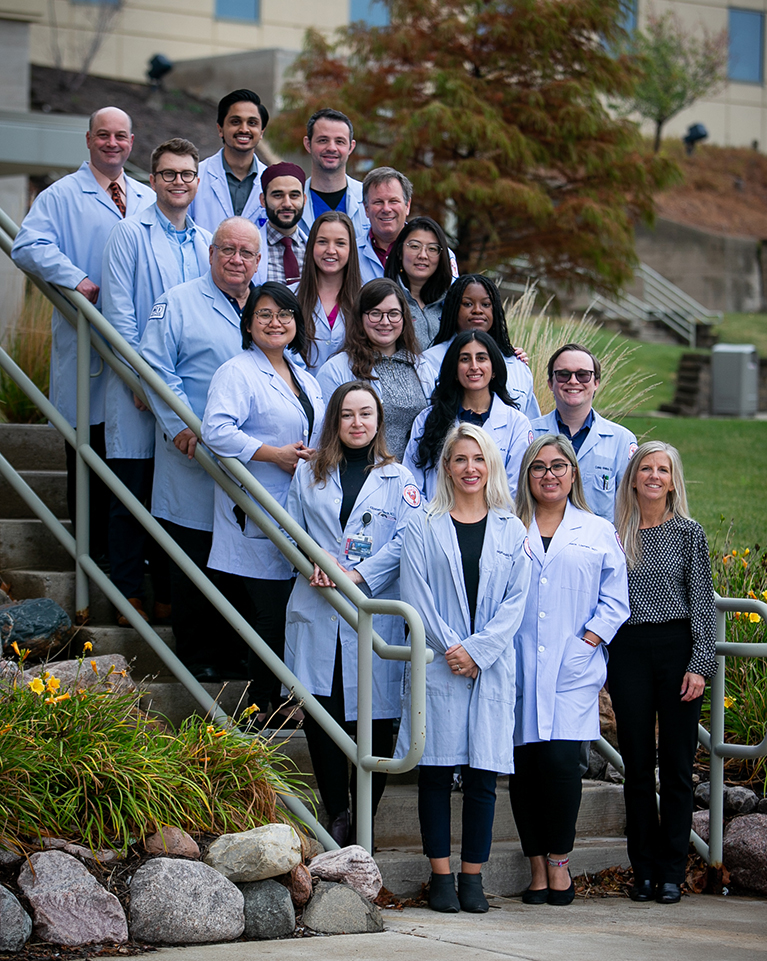 A group of medical students stand on a staircase, smiling and posing in lab coats.
