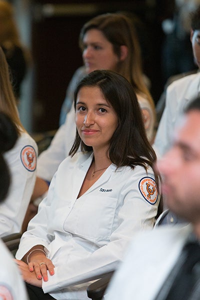 A resident in a white coat smiles while sitting in a row of other residents